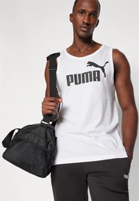 Biely bezrukávový top s čiernym logom "PUMA", kombinovaný s čiernymi nohavicami. Model drží kompaktnú čiernu tašku s sieťovanými detailmi.