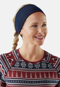 Femme souriante avec des cheveux blonds tressés, portant un bandeau bleu marine et un pulls à motifs rouge, blanc et bleu avec des thèmes d'hiver et de ski.