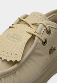 Chaussure bateau en cuir beige avec lacets crème, étiquette logo embossée et accents texturés. Comprend des œillets en métal et un emblème de crocodile.