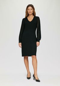 Robe noire avec une silhouette ajustée, décolleté en V, manches longues en dentelle rayée, et une longueur de genoux. Assortie avec des talons noirs pointus.