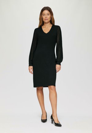 Robe noire avec une silhouette ajustée, décolleté en V, manches longues en dentelle rayée, et une longueur de genoux. Assortie avec des talons noirs pointus.
