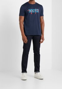 T-shirt di cotone navy con testo grafico rosso e turchese, abbinata a jeans scuri e sneakers bianche. Design semplice e informale.