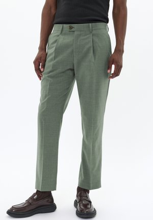 Pantalones ajustados verde claro con una textura sutil, pliegues delanteros y cierre de botón, combinados con mocasines marrón oscuro.