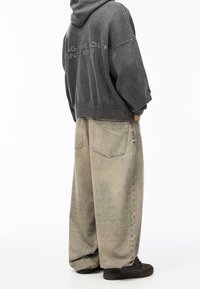 Personne portant un jean beige surdimensionné et décoloré, un sweat à capuche gris foncé avec du texte dans le dos, et des baskets noires, se tenant avec les mains dans les poches.