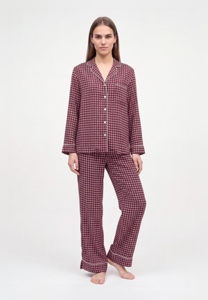Une femme porte un ensemble de pyjama bordeaux à motifs blancs, boutonné, à manches longues et avec un pantalon assorti, sur un fond blanc uni.