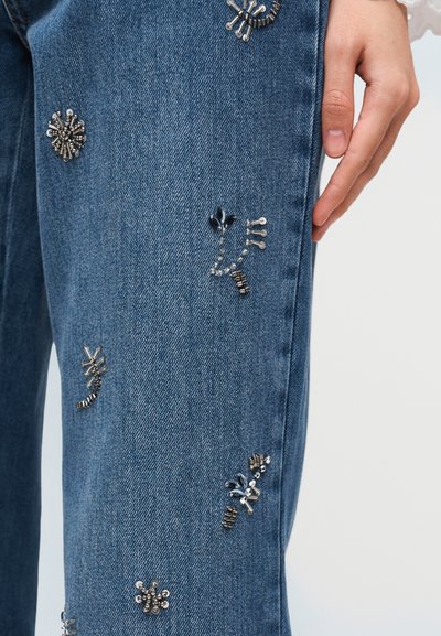 Φωτεινό γαλάζιο τζιν με ασημένιες μεταλλικές διακοσμήσεις και λουλουδάτα μοτίβα, με ίσια γραμμή και ελαφριά υφή.