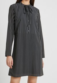 Abito nero con righe verticali bianche e blu, maniche lunghe, dettaglio al collo con fiocco e chiusura a bottoni. Tessuto leggero e vestibilità ampia.