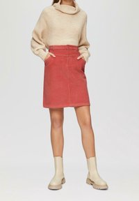 Jupe en velours côtelé de couleur rouille avec une fermeture à boutons au dos et deux poches latérales, associée à un pull en maille oversize beige et des bottines crème.