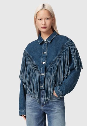 Ung person med langt blondt hår iført en blå cowboyjakke med lange frynser på brystet og ærmerne.