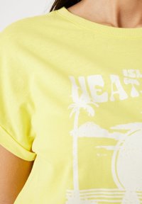 Gul bomulls T-shirt med kort ärm, med ett vitt grafiskt tryck av palmträd och en solmot if. Mjuk känsla, rundad halsdesign.