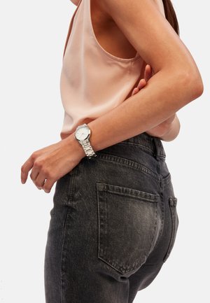 Montre en argent au poignet avec un cadran rond, posée sur un débardeur rose clair et un jean taille haute gris foncé, mettant en valeur la texture et le design.