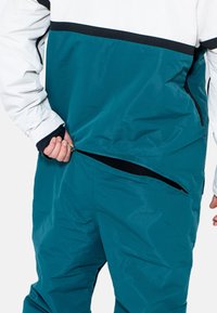 Tannengrüner und weißer Pullover mit schwarzen Akzenten; der Stoff ist glatt und glänzend. Verfügt über eine seitliche Reißverschlusstasche und einen lockeren Schnitt.