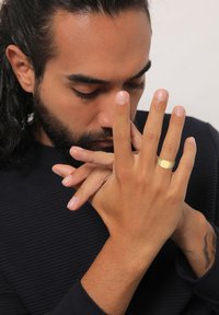 Gold-toned ring with a textured surface worn on a hand. Close-up of fingers and skin tone against a dark shirt.