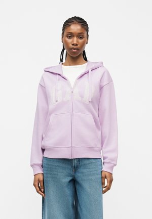 Femme aux cheveux tressés portant un sweat à capuche léger violet clair avec le texte "GAP" et un jean bleu ample, debout devant un fond uni.