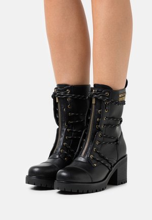 Mustat nahkaiset keskipitkät saappaat, joissa on paksut korot, etuvetoketju, mustavalkoiset nauhat ja kultainen logokilpi ulkosivulla.