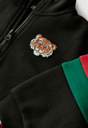 Černá mikina s kapucí na zip s vyšitým designem oranžov�ého a bílého tygra na hrudi a barevnými pruhovanými akcenty na rukávech.