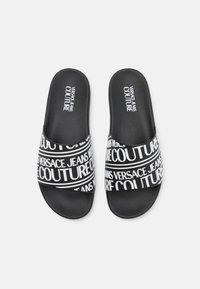 Fekete csúszásmentes papucsok texturált talppal, a pántokon feltűnő fehér nyomtatott "VERSACE JEANS COUTURE" felirattal.
