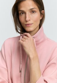 Svetloružová bunda na zips s vysokým golierom, s hladkou textúrou a striebornými akcentmi, ktorú nosí modelka s hnedými vlasmi.