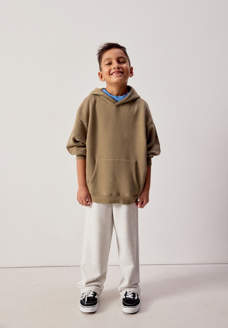 Uśmiechający się chłopiec stojący, ubrany w brązową, za dużą bluzę z kapturem, jasnoszare spodnie dresowe oraz czarno-białe trampki, na jednolitym tle.
