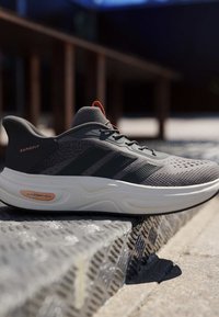 Grauer und schwarzer Sportschuh mit weißer, gepolsterter Sohle mit der Aufschrift „Cloudfoam Cushion“, der auf einer metallisch strukturierten Oberfläche im Freien liegt.