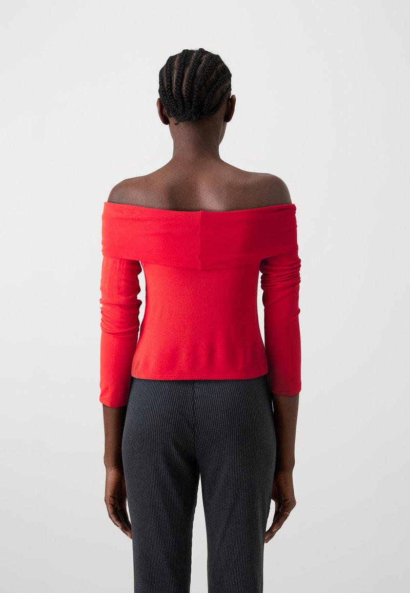 Rotes schulterfreies Oberteil mit langen Ärmeln, aus dehnbarem Stoff gefertigt. Hat ein figurbetontes Design und einen verkürzten Saum. Kombiniert mit grauen Ripp-Hosen.