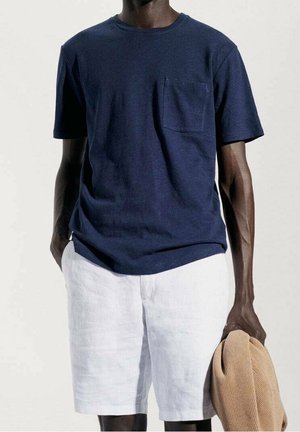 Navyblaues Baumwoll-T-Shirt mit kurzen Ärmeln und Brusttasche, kombiniert mit weißen Leinen-Shorts. Hellbrauner Pullover über dem Arm getragen.