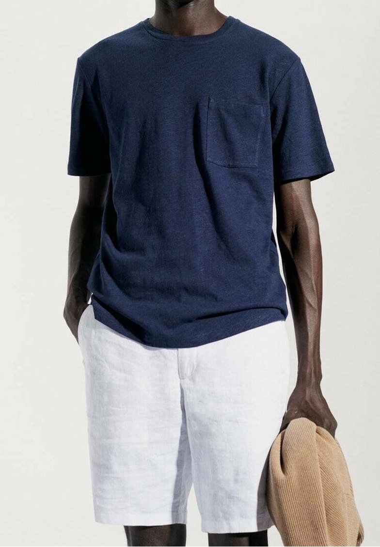 T-shirt en coton bleu marine à manches courtes avec une poche sur la poitrine, associé à un short en lin blanc. Un pull beige clair est drapé sur le bras.