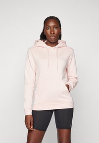 Hoodie rosa claro com um bolso frontal, cordões e um pequeno logo. Combinado com calções pretos, mostrando uma textura suave e um ajuste casual.