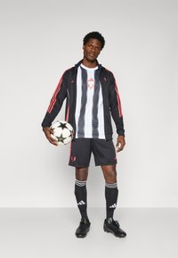 Black sports jacket with red stripes, white and black striped jersey, black shorts, and black soccer cleats holding a soccer ball.