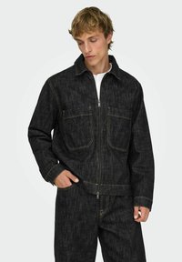 Mustast teksajope lukuga ees, kahe rinnatasku ja kontrastse kollase õmblusega. Silmapaistev tekstuur ja lõdvestatud lõige.