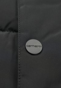 Svart puffjacka av tyg med horisontell söm, med en rund svart knapp med "carhartt" präglat på ytan.