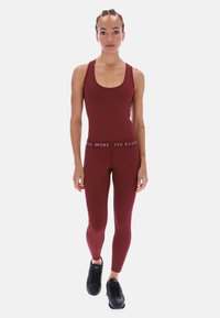 Bordó sportos overall mély kivágással, testhez simuló fazonnal és márkázott derékpánttal. Fekete sportcipővel párosítva.