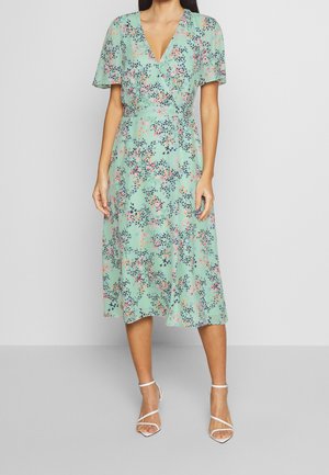 Femme portant une robe midi fleurie vert menthe avec des manches volantées et des sandales blanches à talons avec des brides, debout devant un fond uni.
