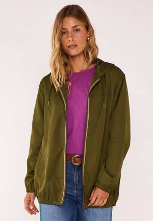 Frau trägt eine olivgrüne Kapuzenjacke mit Reißverschluss über einem lila Hemd, kombiniert mit blauen Jeans und einem braunen Gürtel mit strukturiertem Schnallenverschluss.