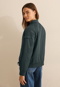 Giacca bomber verde scuro con colletto e polsini a coste, dotata di tasche laterali e cordoncino regolabile all'orlo. Indossata con jeans blu.