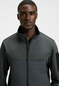 Mörkgrå zip-up-jacka med en strukturerad övre del, slätt nedre tyg och en dragkedjeficka på vänster arm. Designad för funktionalitet.