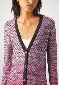 Femme portant un cardigan en maille texturée violet et rose avec des bordures noires et des boutons roses, par-dessus un haut blanc, visible des épaules à la taille.