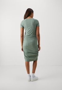 Grünes geripptes Kleid mit kurzen Ärmeln, figurbetonter Silhouette und Raffung an der Seite. Das Material wirkt weich mit leichtem Glanz.
