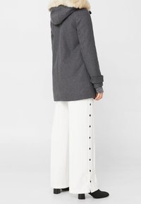 Manteau en laine grise avec une capuche en fourrure, présentant une coupe droite et des détails de boutons sur un pantalon large blanc. Des bottes à talons noires complètent la tenue.