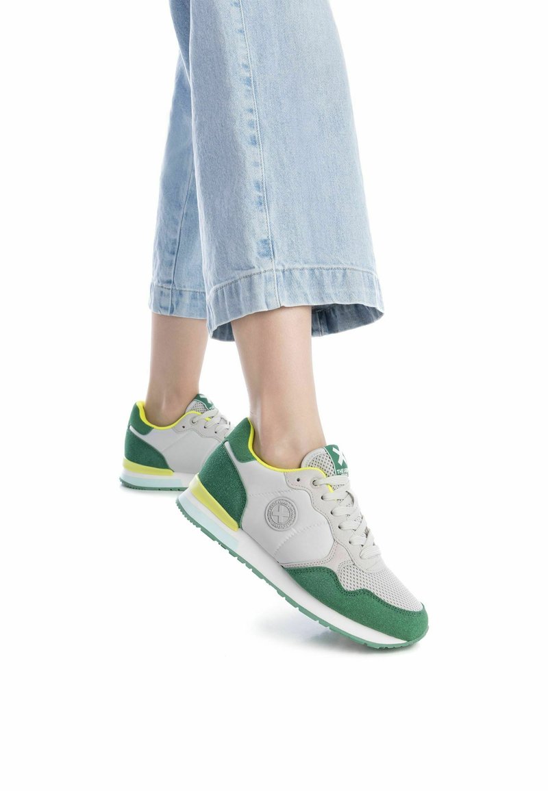 Zapatillas atléticas en gris y verde con materiales de malla y ante, que presentan un acento amarillo y un logo, combinadas con unos jeans de pierna ancha en azul claro.