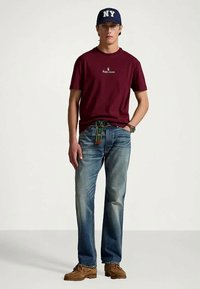 Ung man som bär en vinröd Ralph Lauren t-shirt, blå jeans med tygbälte, bruna skor, runda glasögon och en marinblå NY-baseballkeps.