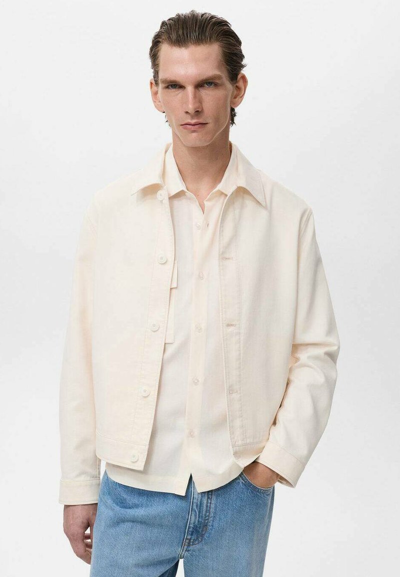 Uomo con capelli all'indietro, che indossa una camicia crema con bottoni e una giacca abbinata, abbinata a jeans azzurri chiari, pose davanti a uno sfondo semplice.