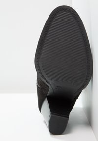 Podeszwa i obcas czarnego zamszowego buta na obcasie opierającego się o białą powierzchnię, pokazujące teksturowaną gumową podeszwę i masywny obcas.