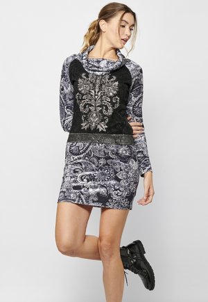 Robe à manches longues avec un corsage en velours noir ornée de broderies argentées intriquées, associée à une jupe à motif paisley, stylée avec des bottes noires.