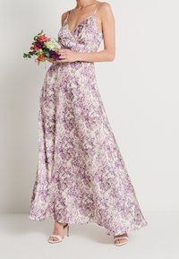 Robe longue fleurie avec un décolleté en V et des bretelles fines, présentant un imprimé botanique violet et vert sur un fond crème. Portée avec des talons à brides.