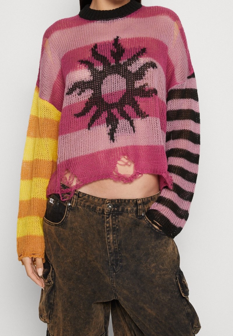 Femme portant un pull court en maille rose avec un motif soleil noir, bords effilochés, manches rayées dépareillées et pantalon cargo marron.