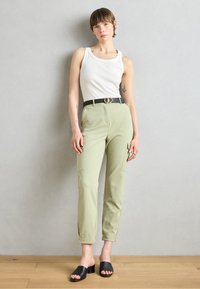 Pantaloni cargo verzi deschisi cu buzunare laterale, talie ajustată și o curea neagră cu cataramă dublă circulară. Purtați cu un maiou alb și papuci negri.
