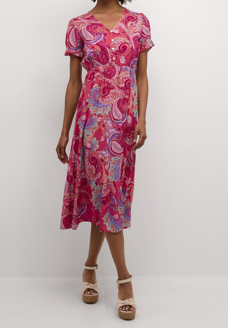 Femme portant une robe midi rose avec des motifs cachemire multicolores, des manches courtes bouffantes, des boutons sur la poitrine et des sandales à plateforme beige.