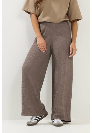 Osoba nosząca teksturowane szerokie brązowe spodnie, beżowy t-shirt i białe trampki z czarnymi paskami na jednolitym tle.