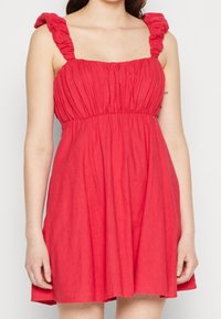 Robe rouge sans manches avec un corsage froncé et des bretelles à volants. Silhouette en A, longueur genou, fabriquée à partir d'un tissu texturé.